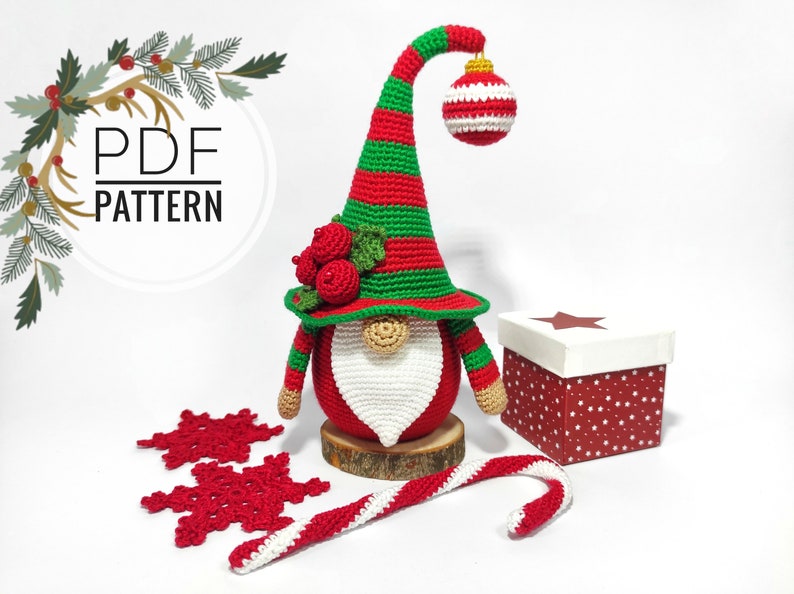 May include: A handmade crochet Christmas gnome with a red and green striped hat, a white beard, and a red body. The gnome is decorated with red berries and a hanging ornament. Includes a candy cane, snowflakes, and a gift box.
