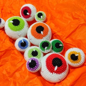 K&ouml;nnte beinhalten: Eine Sammlung von neun geh&auml;kelten Aug&auml;pfeln in verschiedenen Farben, darunter Rot, Orange, Gr&uuml;n, Blau und Lila. Die Aug&auml;pfel sind alle unterschiedlich gro&szlig; und haben wei&szlig;es Garn f&uuml;r die Sklera, mit schwarzem Garn f&uuml;r die Pupillen.