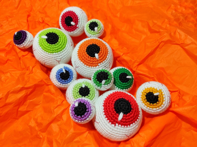 K&ouml;nnte beinhalten: Eine Sammlung von neun geh&auml;kelten Aug&auml;pfeln in verschiedenen Farben, darunter Rot, Orange, Gr&uuml;n, Blau und Lila. Die Aug&auml;pfel sind alle unterschiedlich gro&szlig; und haben wei&szlig;es Garn f&uuml;r die Sklera, mit schwarzem Garn f&uuml;r die Pupillen.