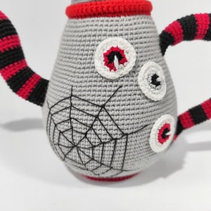 K&ouml;nnte beinhalten: Eine graue geh&auml;kelte Teekanne mit einem Spinnennetz-Design und drei wei&szlig;en geh&auml;kelten Augen. Die Teekanne hat einen schwarz-rot gestreiften Griff.