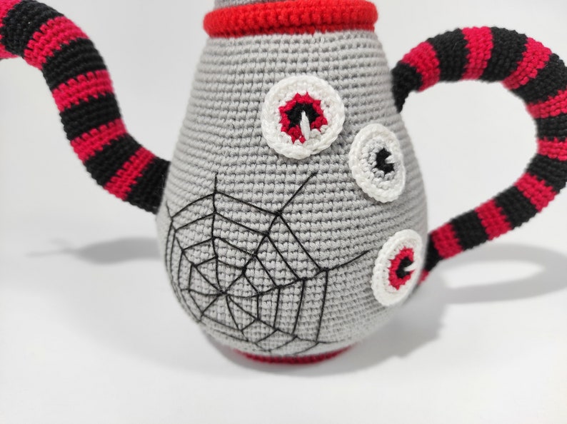 K&ouml;nnte beinhalten: Eine graue geh&auml;kelte Teekanne mit einem Spinnennetz-Design und drei wei&szlig;en geh&auml;kelten Augen. Die Teekanne hat einen schwarz-rot gestreiften Griff.