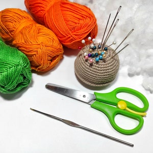 Puede incluir: Primer plano de un alfiletero de ganchillo con alfileres de colores, ovillos de lana verde, naranja y blanca, un par de tijeras verdes y un ganchillo sobre una superficie blanca.
