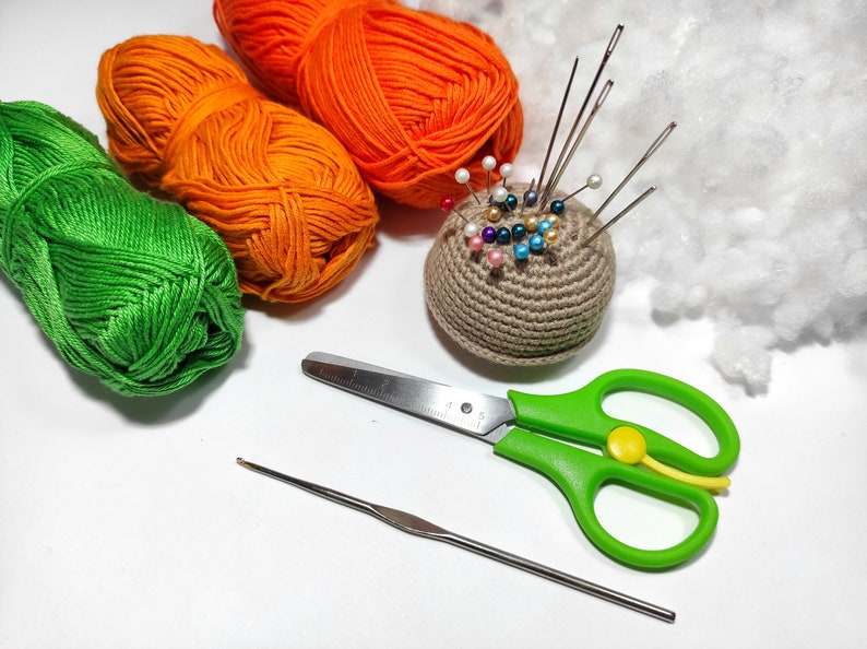 Puede incluir: Primer plano de un alfiletero de ganchillo con alfileres de colores, ovillos de lana verde, naranja y blanca, un par de tijeras verdes y un ganchillo sobre una superficie blanca.