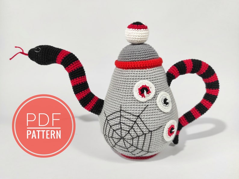 K&ouml;nnte beinhalten: Eine geh&auml;kelte Teekanne mit einem Schlangenhenkel, einem Spinnennetz-Design und drei Augen. Die Teekanne ist grau mit roten und schwarzen Streifen am Henkel. Die Teekanne ist ein PDF-Muster.