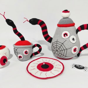 K&ouml;nnte beinhalten: Ein geh&auml;keltes Teeset mit Teekanne, Tasse, Untertasse und zwei Augen. Die Teekanne und die Tasse sind mit roten und schwarzen Streifen und einem Spinnennetz-Design verziert. Die Untertasse ist mit einem gro&szlig;en Auge verziert.
