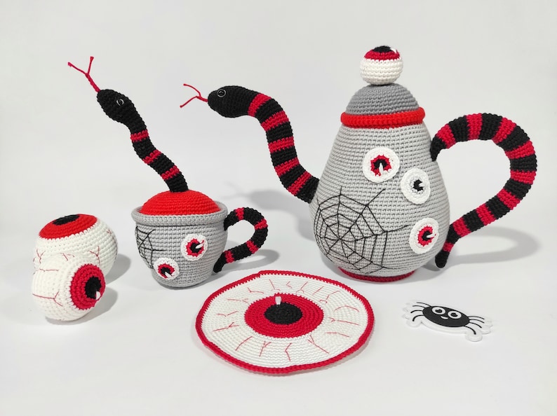 K&ouml;nnte beinhalten: Ein geh&auml;keltes Teeset mit Teekanne, Tasse, Untertasse und zwei Augen. Die Teekanne und die Tasse sind mit roten und schwarzen Streifen und einem Spinnennetz-Design verziert. Die Untertasse ist mit einem gro&szlig;en Auge verziert.