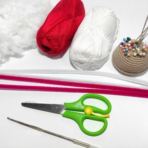 May include: A collection of craft supplies, including red and white yarn balls, white stuffing, pink and white pipe cleaners, a crochet hook, green scissors, and a brown pincushion with colorful pins. Perfect for crafting projects.