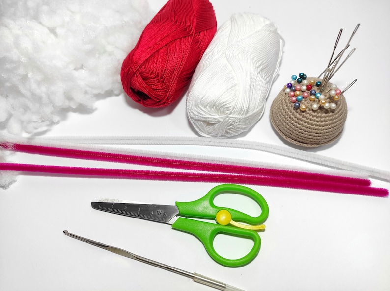 May include: A collection of craft supplies, including red and white yarn balls, white stuffing, pink and white pipe cleaners, a crochet hook, green scissors, and a brown pincushion with colorful pins. Perfect for crafting projects.