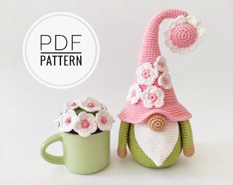 May include: A pink crocheted gnome with a white beard and green body. The gnome is wearing a pink hat with white flowers and a pink flower on top. The gnome is sitting on a white surface.  PDF PATTERN is written on a white circle.