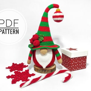 May include: A crocheted Christmas gnome with a red body, white beard, and a green and red striped hat. The hat has a hanging ornament. Includes a gift box, snowflakes, and a candy cane. The text "PDF PATTERN" is in a circle.