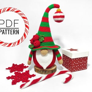 May include: A handmade crochet Christmas gnome with a red and green striped hat, a white beard, and a red body. The gnome is decorated with a red ornament and berries. Includes a candy cane, snowflakes, and a gift box. The image also includes the text "PDF PATTERN".