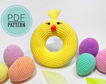 May include: Crochet pattern for a yellow chick ring toy with black eyes and a red nose. The pattern also includes instructions for making smaller crocheted eggs in various colors, including purple, pink, and green.