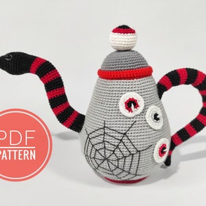 Puede incluir: Tetera de amigurumi de crochet con un asa de serpiente a rayas negras y rojas, cuerpo gris y ojos blancos. La tetera tiene un diseño de telaraña y un borde rojo.  PATRÓN PDF.