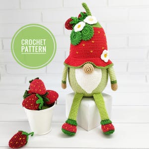 May include: A crocheted gnome with a red strawberry hat and green body. The gnome has a white heart-shaped belly and is sitting on a white block. A bucket of crocheted strawberries is next to the gnome. The image includes the text "CROCHET PATTERN".
