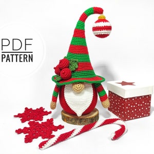 May include: A handmade crochet Christmas gnome with a red body, white beard, and a green and red striped hat. The gnome holds a red and white striped ornament. The image also includes a gift box, snowflakes, and a candy cane.