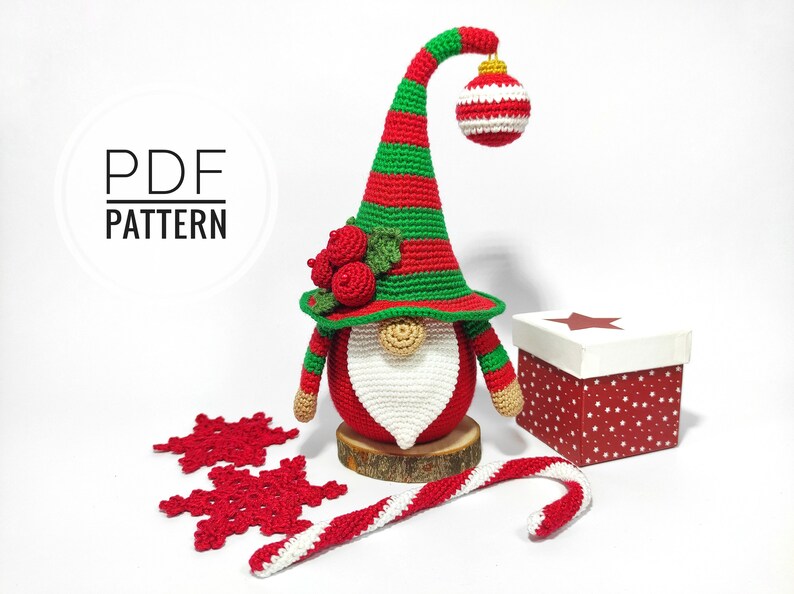 May include: A handmade crochet Christmas gnome with a red body, white beard, and a green and red striped hat. The gnome holds a red and white striped ornament. The image also includes a gift box, snowflakes, and a candy cane.