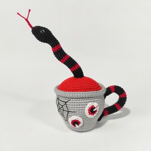 K&ouml;nnte beinhalten: Eine geh&auml;kelte Tasse mit einer rot-schwarzen Schlange, die aus der Oberseite ragt. Die Tasse hat ein Spinnennetz-Design und zwei gro&szlig;e Augen auf der Vorderseite.