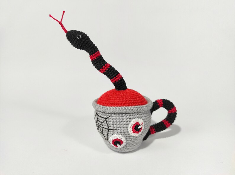 K&ouml;nnte beinhalten: Eine geh&auml;kelte Tasse mit einer rot-schwarzen Schlange, die aus der Oberseite ragt. Die Tasse hat ein Spinnennetz-Design und zwei gro&szlig;e Augen auf der Vorderseite.
