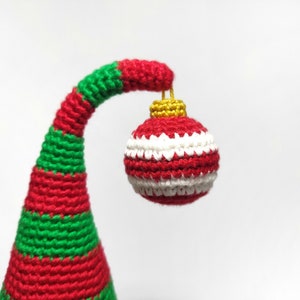 May include: A handmade Christmas ornament, featuring a red, white, and green color scheme. The ornament is a crochet design, with a red and white striped ball suspended from a curved, striped stem. The stem is red and green.