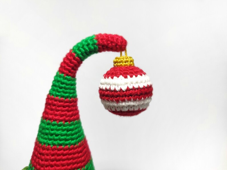 May include: A handmade Christmas ornament, featuring a red, white, and green color scheme. The ornament is a crochet design, with a red and white striped ball suspended from a curved, striped stem. The stem is red and green.