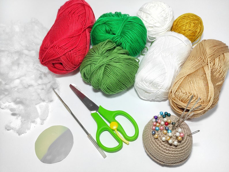 May include: A collection of craft supplies for crocheting or knitting, including red, green, white, and tan yarn balls, stuffing, a crochet hook, scissors, a pin cushion with pins, and a small, two-tone disc.