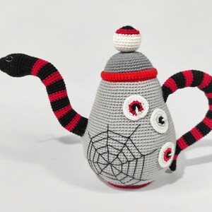 K&ouml;nnte beinhalten: Eine graue geh&auml;kelte Teekanne mit einem schwarz-rot gestreiften Schlangenhenkel und einem wei&szlig;en Auge oben. Die Teekanne hat ein Spinnennetz-Design und drei wei&szlig;e Augen auf der Vorderseite.