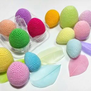 Puede incluir: Un conjunto de huevos de Pascua de ganchillo en varios colores pastel, incluyendo rosa, amarillo, verde, azul y morado. Los huevos est&aacute;n dispuestos en un recipiente de pl&aacute;stico transparente y sobre una superficie blanca.