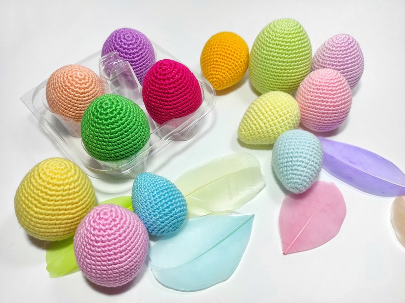 Puede incluir: Un conjunto de huevos de Pascua de ganchillo en varios colores pastel, incluyendo rosa, amarillo, verde, azul y morado. Los huevos est&aacute;n dispuestos en un recipiente de pl&aacute;stico transparente y sobre una superficie blanca.