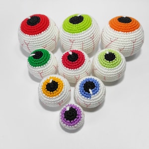 K&ouml;nnte beinhalten: Ein Set aus neun geh&auml;kelten Aug&auml;pfeln in verschiedenen Farben, darunter Rot, Gr&uuml;n, Orange, Gelb, Blau und Lila. Die Aug&auml;pfel sind alle wei&szlig; mit einer schwarzen Pupille und einer unterschiedlich gef&auml;rbten Iris.