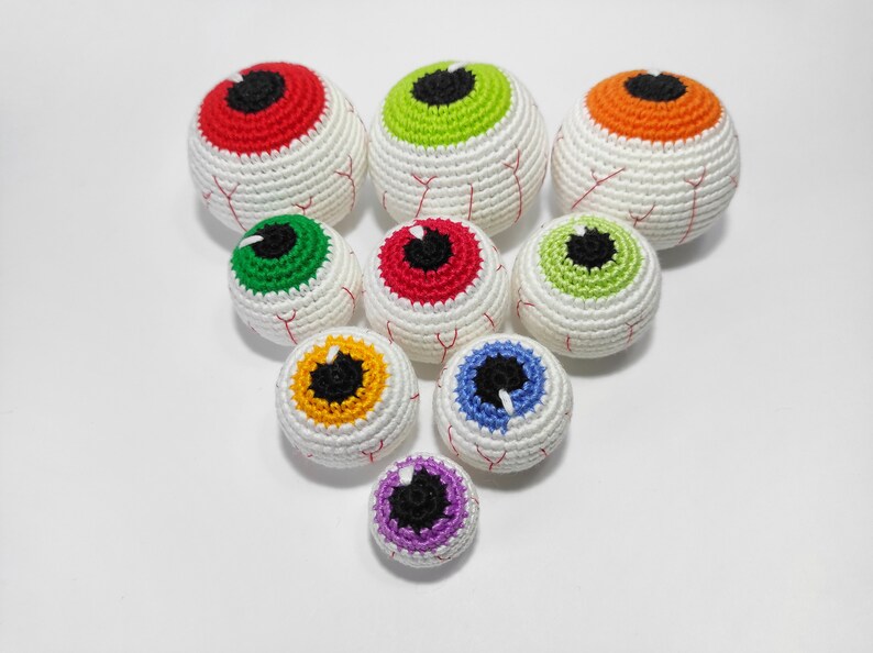 K&ouml;nnte beinhalten: Ein Set aus neun geh&auml;kelten Aug&auml;pfeln in verschiedenen Farben, darunter Rot, Gr&uuml;n, Orange, Gelb, Blau und Lila. Die Aug&auml;pfel sind alle wei&szlig; mit einer schwarzen Pupille und einer unterschiedlich gef&auml;rbten Iris.