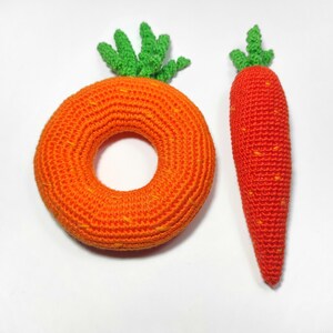 Puede incluir: Dos zanahorias de crochet, una es un anillo y la otra es una zanahoria larga. Ambas son de color naranja con puntas verdes.