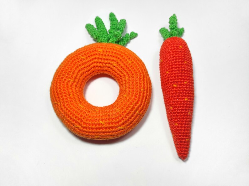 Puede incluir: Dos zanahorias de crochet, una es un anillo y la otra es una zanahoria larga. Ambas son de color naranja con puntas verdes.