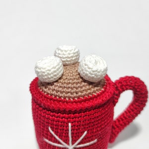 May include: A crocheted mug with a red exterior, brown interior, and white marshmallows on top. The mug has a snowflake design on the front.