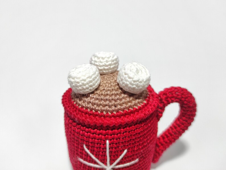 May include: A crocheted mug with a red exterior, brown interior, and white marshmallows on top. The mug has a snowflake design on the front.