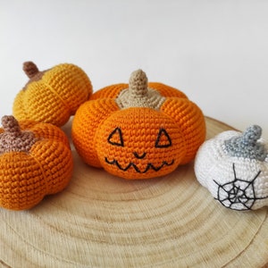 May include: Four crocheted pumpkins on a wooden surface. One pumpkin is white with a black spiderweb, and the other three are orange with black facial features.