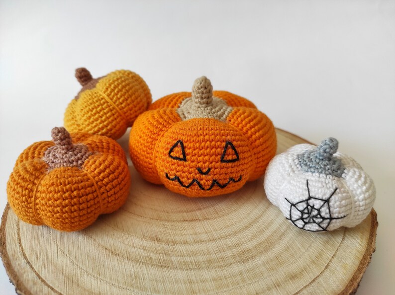 May include: Four crocheted pumpkins on a wooden surface. One pumpkin is white with a black spiderweb, and the other three are orange with black facial features.