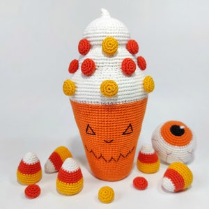 May include: A crocheted Halloween-themed candy corn with a white top, orange bottom, and yellow and red candy dots. There are also several smaller crocheted candy corn and a crocheted eyeball.