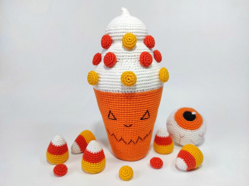 May include: A crocheted Halloween-themed candy corn with a white top, orange bottom, and yellow and red candy dots. There are also several smaller crocheted candy corn and a crocheted eyeball.