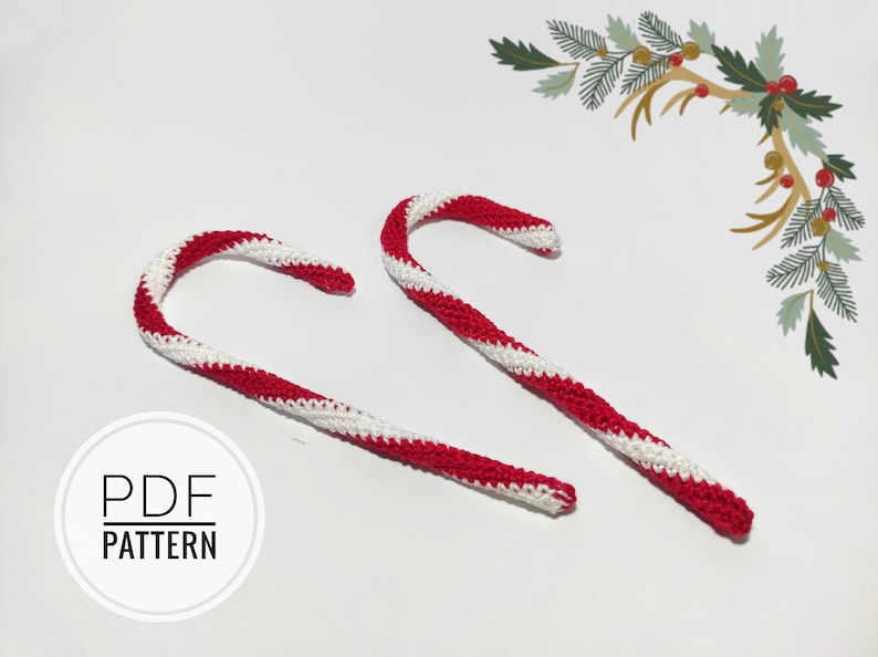 May include: Two handmade crochet candy canes with red and white stripes. The candy canes are curved and have a textured appearance. The image also includes a decorative floral element and the text "PDF PATTERN".