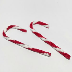 May include: Two handmade crochet candy canes with red and white stripes. The candy canes are curved and have a textured appearance, suggesting a soft, yarn-like material. The background is plain white.