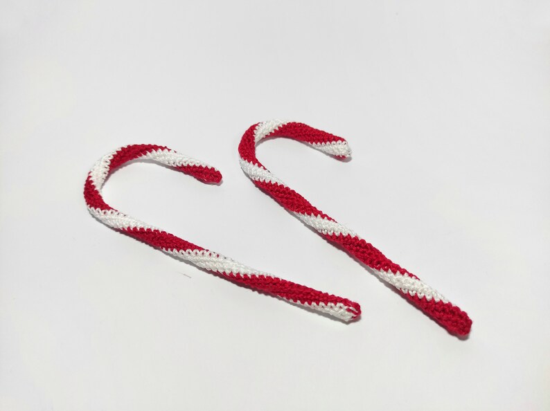 May include: Two handmade crochet candy canes with red and white stripes. The candy canes are curved and have a textured appearance, suggesting a soft, yarn-like material. The background is plain white.