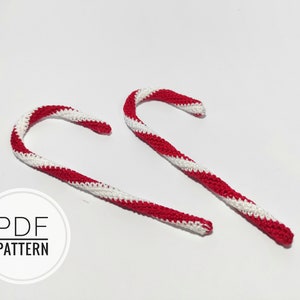 May include: Two crocheted candy canes, one red and white and one white and red, are lying on a white surface. The text "PDF PATTERN" is printed on a white circle.