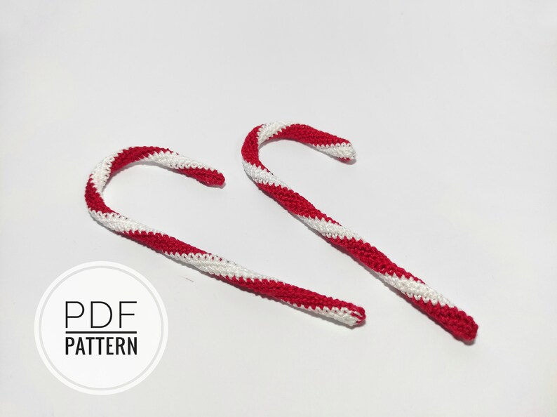 May include: Two crocheted candy canes, one red and white and one white and red, are lying on a white surface. The text "PDF PATTERN" is printed on a white circle.