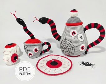 May include: A crocheted tea set featuring a teapot, cup, saucer, and spider. The teapot and cup are decorated with eyes and a spider's web. The saucer is decorated with an eye. The spider is black and white.