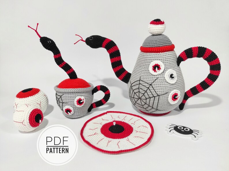 K&ouml;nnte beinhalten: Ein geh&auml;keltes Teeset mit Teekanne, Tasse, Untertasse und Spinne. Die Teekanne und die Tasse sind mit Augen und einem Spinnennetz verziert. Die Untertasse ist mit einem Auge verziert. Die Spinne ist schwarz und wei&szlig;.