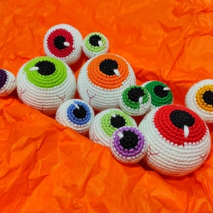 May include: A collection of crocheted eyeball ornaments in various colors, including red, green, orange, yellow, blue, and purple. The eyeballs are arranged on a bright orange surface.