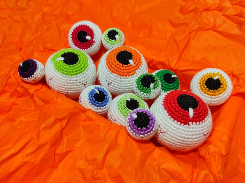 May include: A collection of crocheted eyeball ornaments in various colors, including red, green, orange, yellow, blue, and purple. The eyeballs are arranged on a bright orange surface.