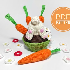Puede incluir: Un cupcake de Pascua tejido a ganchillo con un conejito y zanahorias. El cupcake tiene una base marrón, una cesta verde y un conejito blanco con detalles rosas. Zanahorias naranjas con puntas verdes se colocan alrededor del conejito. La imagen también incluye flores y una zanahoria tejidas a ganchillo.