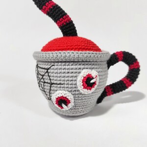 K&ouml;nnte beinhalten: Eine geh&auml;kelte graue Teetasse mit roten und schwarzen Streifen, zwei wei&szlig;en und roten Augen und einem schwarzen Spinnennetz-Design. Die Teetasse hat einen roten und schwarzen gestreiften Griff.