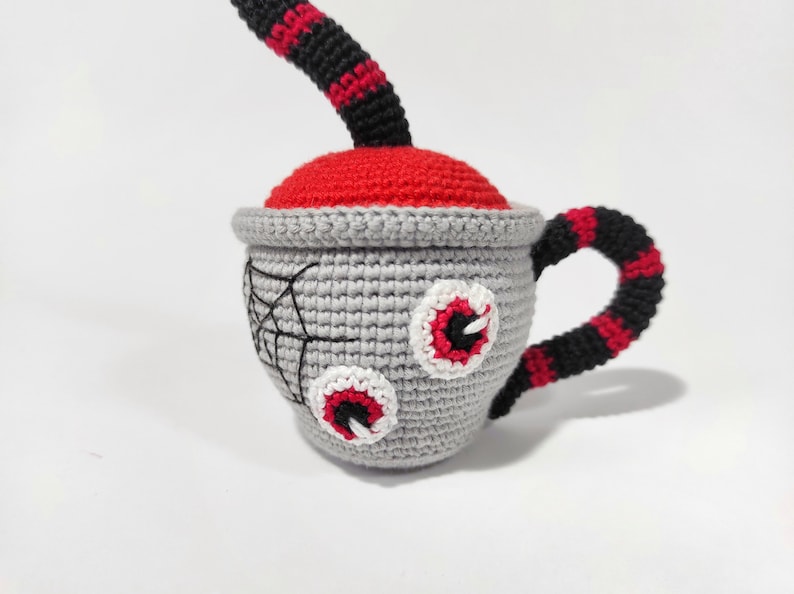 K&ouml;nnte beinhalten: Eine geh&auml;kelte graue Teetasse mit roten und schwarzen Streifen, zwei wei&szlig;en und roten Augen und einem schwarzen Spinnennetz-Design. Die Teetasse hat einen roten und schwarzen gestreiften Griff.